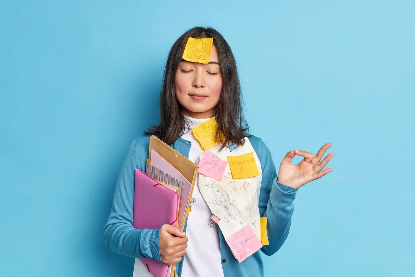 Portrait of a relaxed student who is trying to relax, meditating indoors, making an OK gesture with eyes closed, and holding folders stuffed with papers.