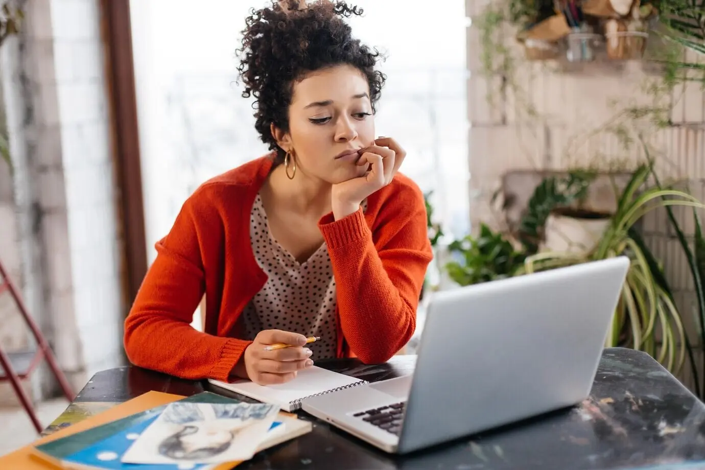 A young, serious woman with dark curly hair is seated at the table, thoughtfully working on a laptop in a modern, cozy workshop with large windows in the background.